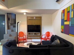 A living room with a black sofa, red chairs, a flat-screen TV, and a colorful abstract painting on the wall. - Loft - Rento (Carapicuíba)