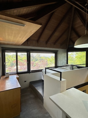 A room with a sloped ceiling, wooden beams, a window with blinds, a bed, a desk, and a hanging light fixture.