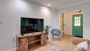 Ett vardagsrum med en stor TV-skärm på ett trästativ, en blå fåtölj, en beige soffa och en grön dörr i bakgrunden.