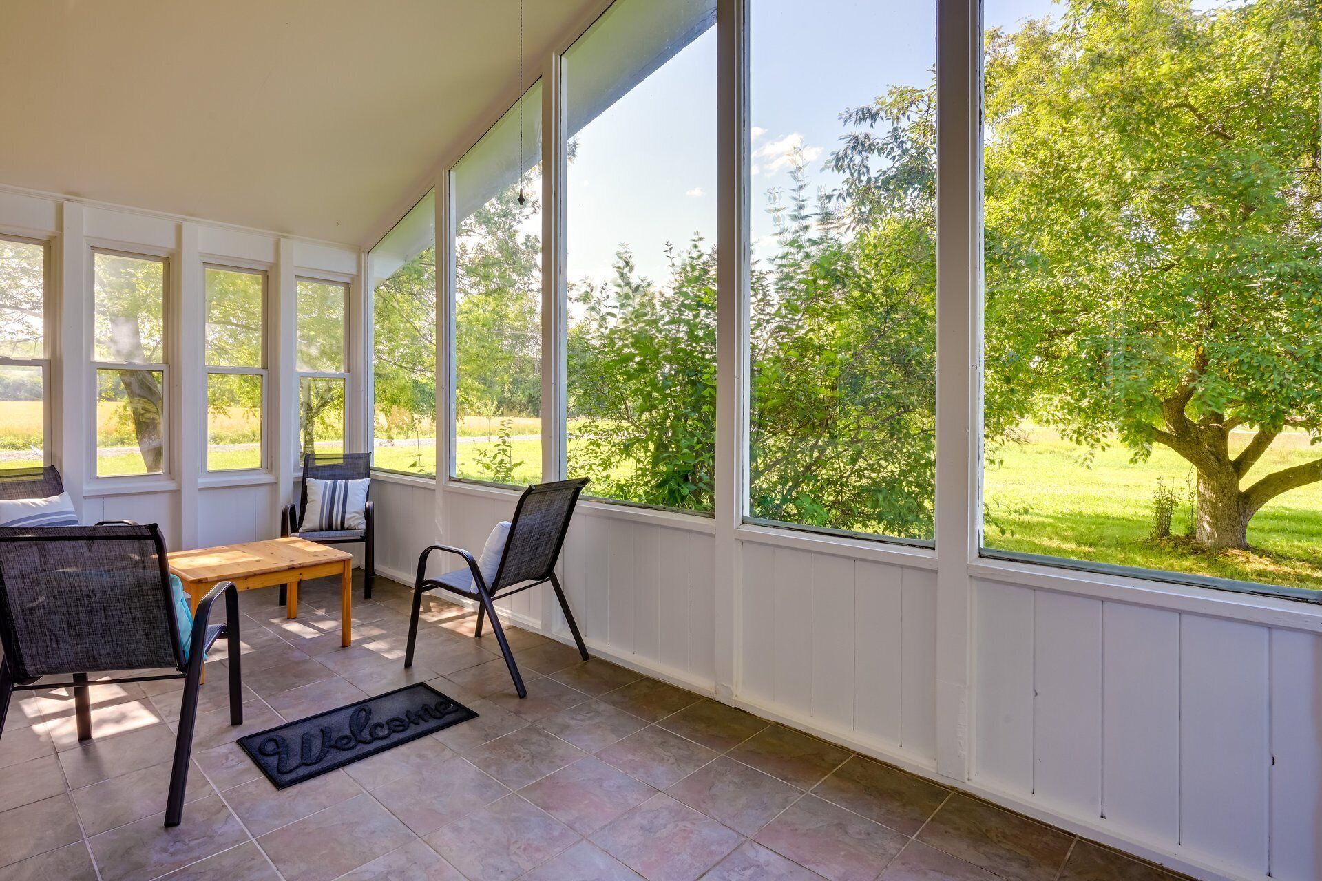 A covered patio with a wooden table and chairs, large windows offering a view of a green landscape.