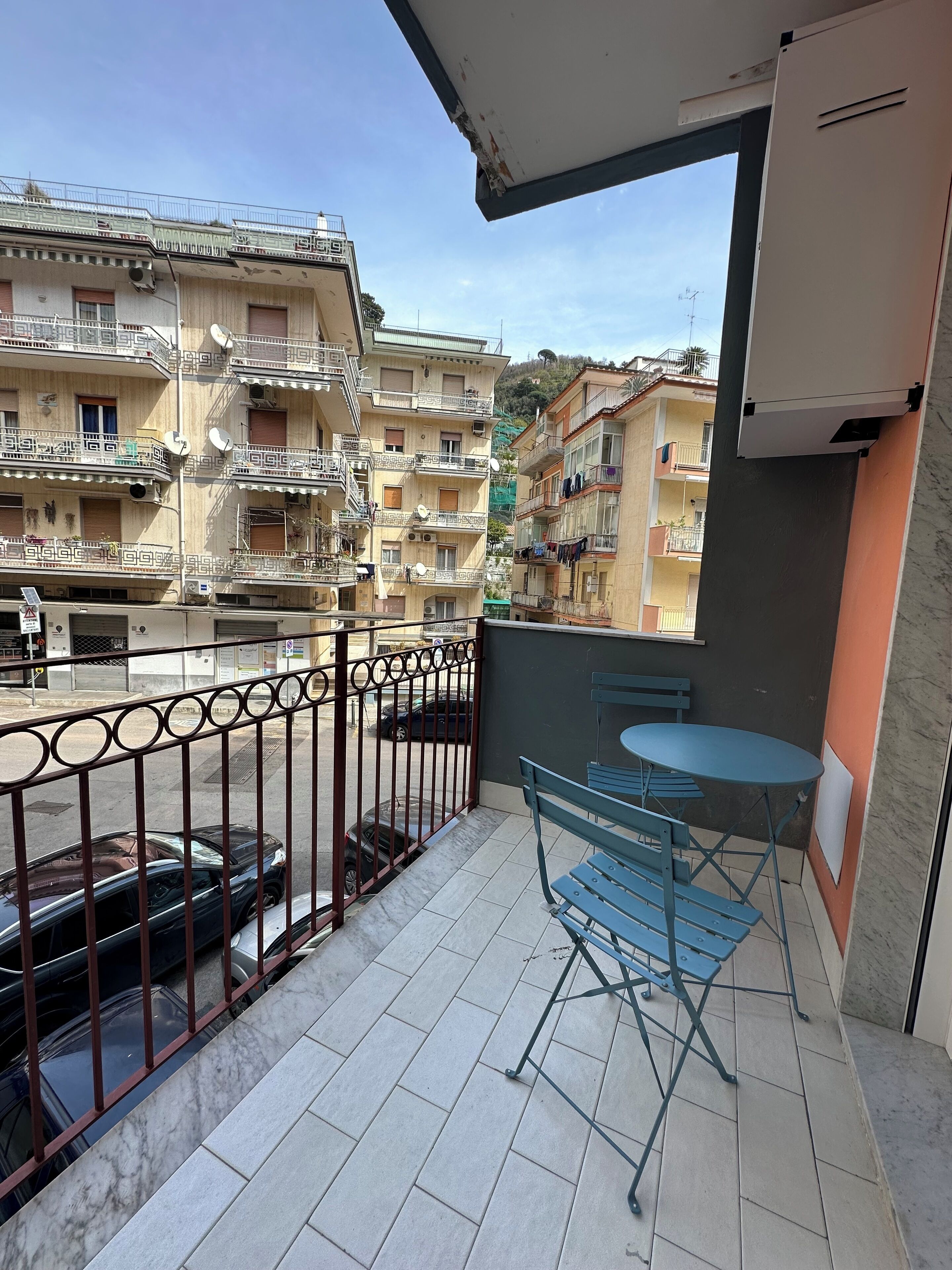 Un balcón con una mesa y sillas azules, barandilla de metal y vistas a edificios de varios pisos.