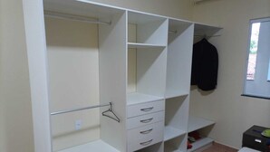 A room with a white wardrobe featuring multiple shelves and drawers, a small stool, and a window with a view of the outside. - House in Guriri for rent (SAO MATEUS)