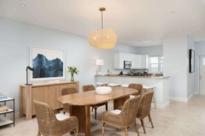 A modern kitchen with white cabinets, a central island, and a dining area with a wooden table and wicker chairs. - 2-Min Walk to Grace Bay Beach Condo (Grace Bay)