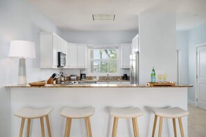 A modern kitchen with white cabinets, a central island with stools, and a window providing natural light. - 2-Min Walk to Grace Bay Beach Condo (Grace Bay)