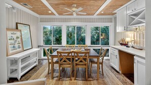A dining area with a wooden table and chairs, large windows with a view of tropical foliage, and a ceiling fan. - Pet Friendly, Tiki Bar, 3 pools, pickleball! (Edisto Island)