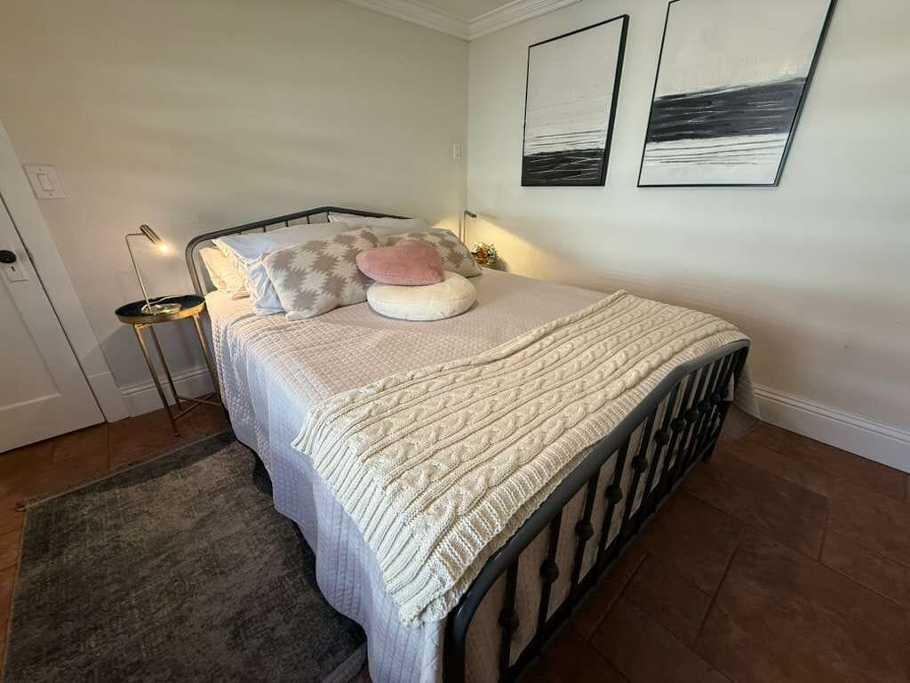 Een net opgemaakt bed met een bedsprei en kussens, een bedlampje en twee ingelijste kunstwerken aan de muur.