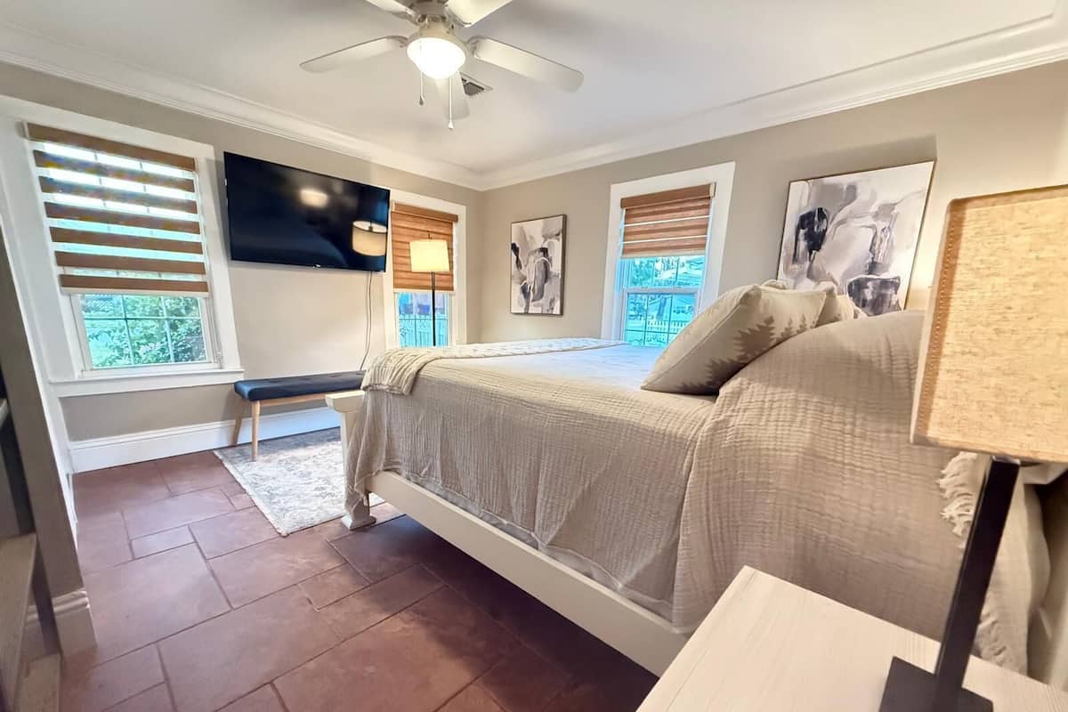 A bedroom with a bed, a ceiling fan, a window with blinds, and artwork on the walls.