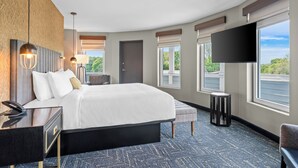 Une chambre d’hôtel moderne dotée d’un grand lit, de tables de chevet, d’un téléviseur à écran plat et de grandes fenêtres offrant une vue sur l’extérieur.