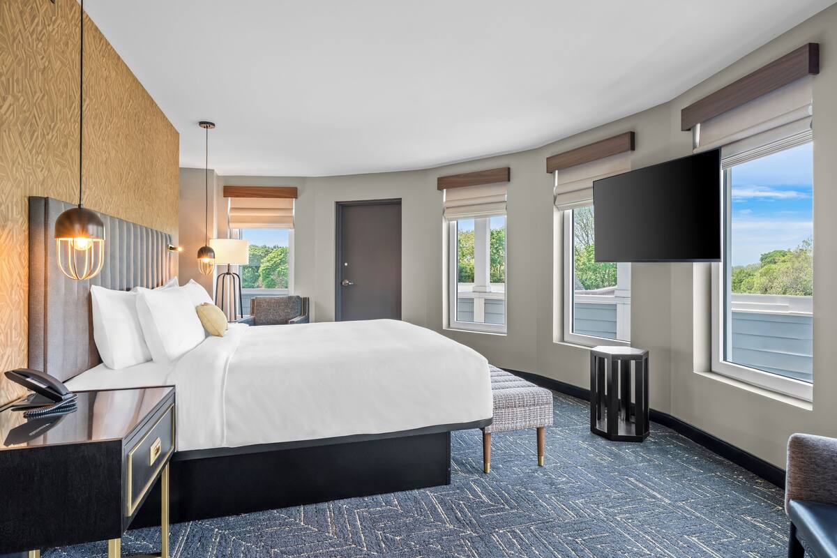 Una habitación de hotel moderna con una cama grande, mesitas de noche, un televisor de pantalla plana y ventanas amplias con vistas al exterior.