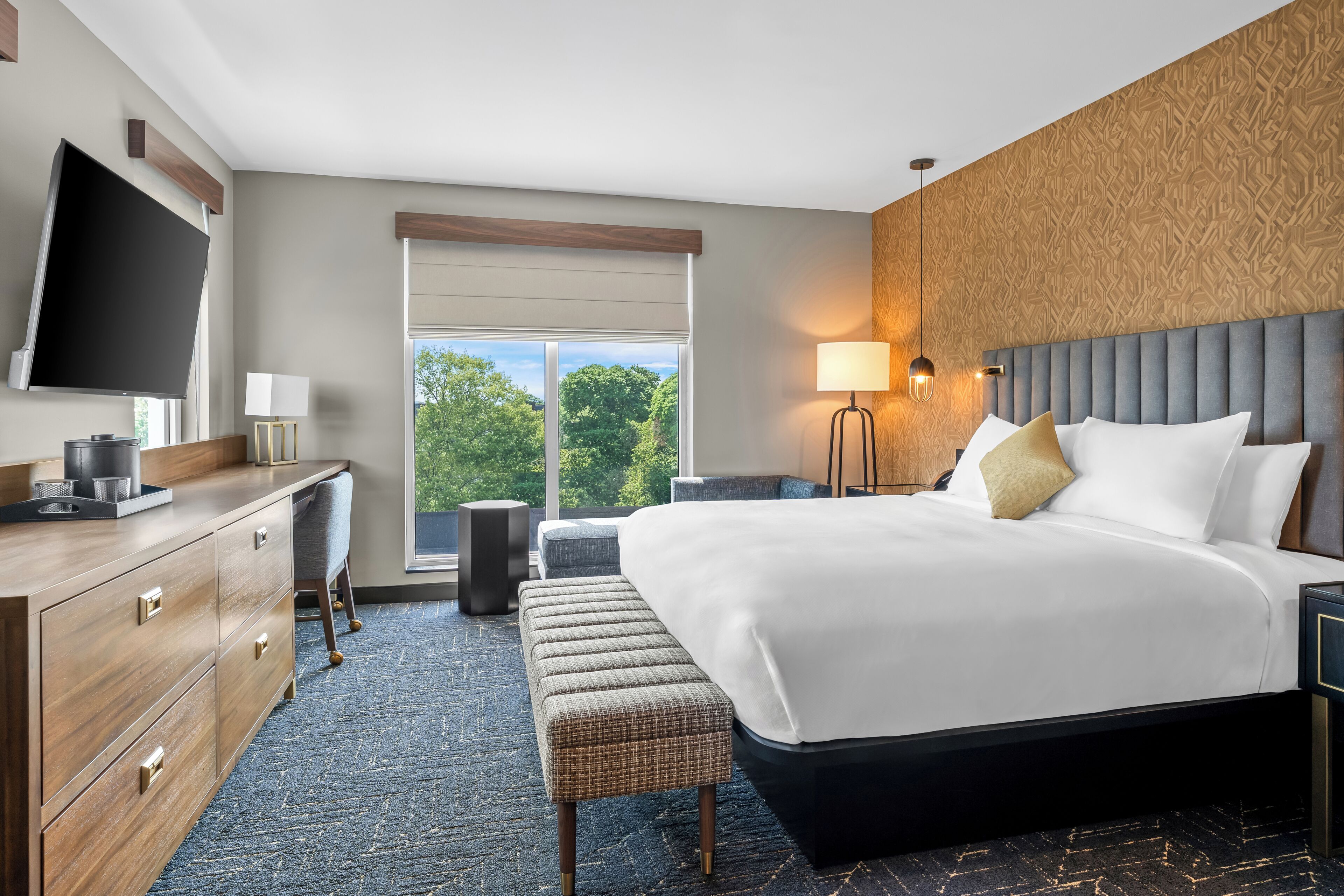 A hotel room with a large bed, a desk with drawers, a flat-screen TV, a bench, and a window with a view of trees.
