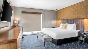 Een hotelkamer met een bed, nachtkastje, bankje, televisie en een raam met jaloezieën.
