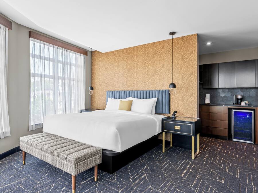 A hotel room with a large bed, bedside tables, a bench, a built-in kitchen area, and a large window.