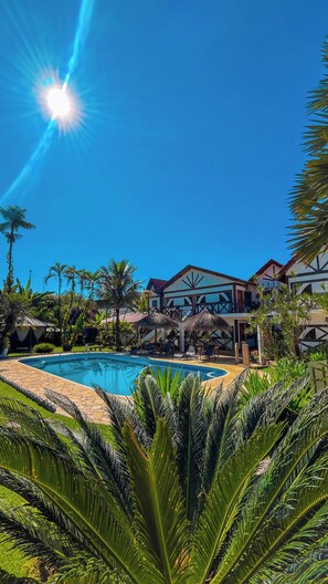 A pool area with a clear blue sky, palm trees, and a building with a balcony. - Pousada Igarapé (Socorro)