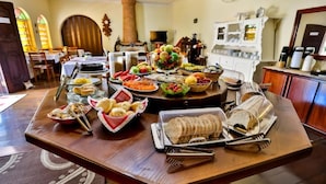 A dining table with various food items and utensils, set in a room with a fireplace, wooden furniture, and a view of the outside through windows. - Pousada Igarapé (Socorro)