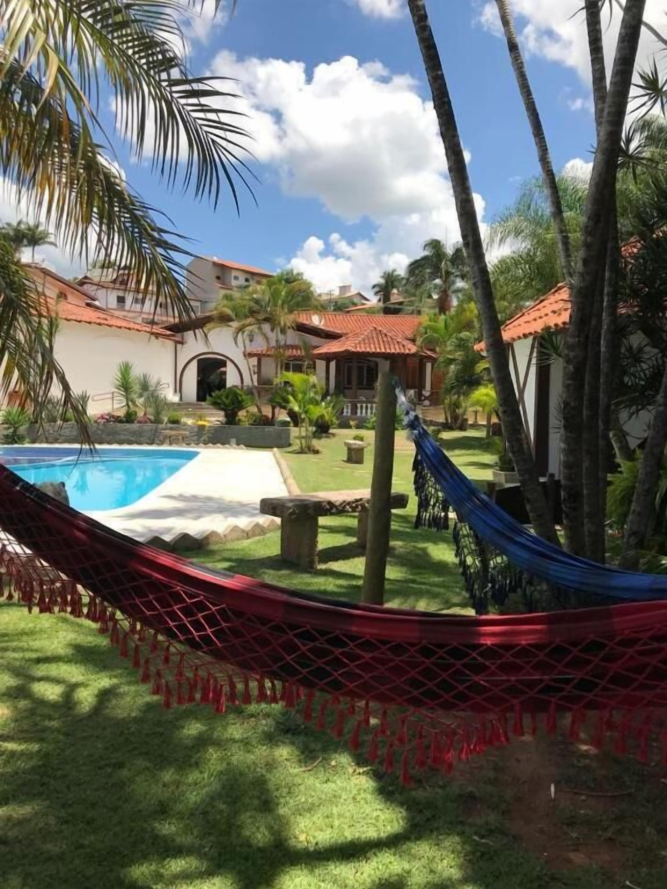 A hammock in a lush garden with a pool and a house with a red-tiled roof.
