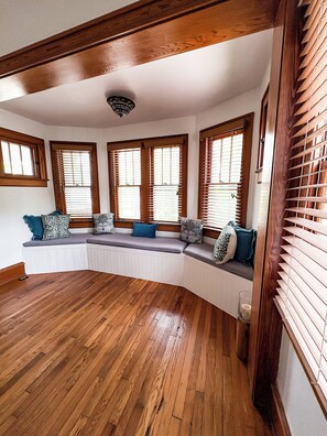 Una habitación con un banco empotrado a lo largo de la pared, suelo de madera y ventanas amplias con persianas.