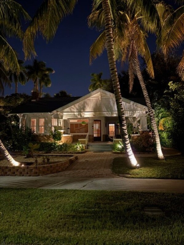 Una casa de dos pisos bien iluminada, con un porche delantero destacado y un techo triangular, rodeada de palmeras y un jardín ajardinado.