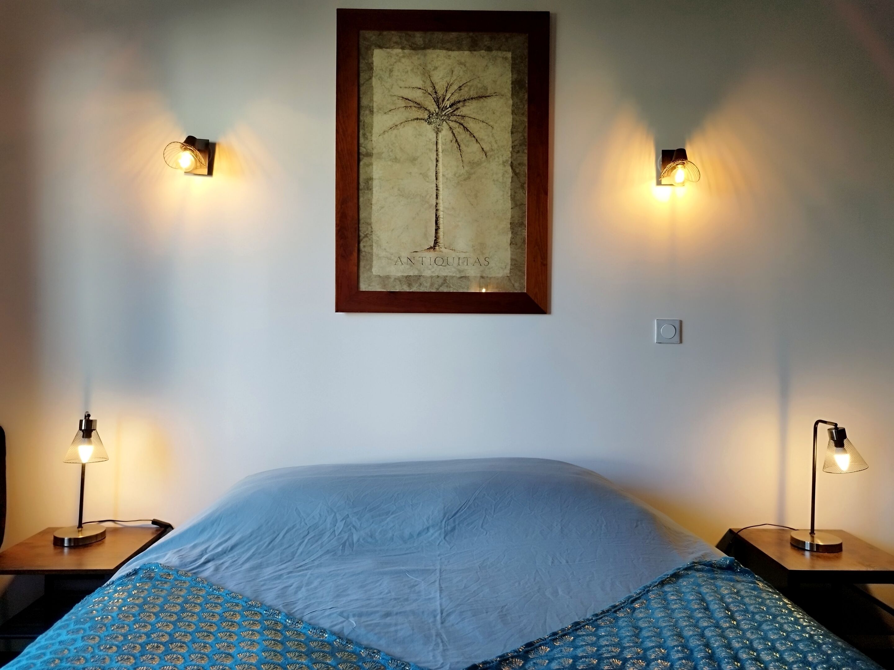A bedroom with a bed, two bedside lamps, a wall-mounted picture frame, and a light switch.