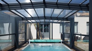 Una piscina cubierta al aire libre con muelle de madera, paredes de vidrio y estructura de techo metálico.