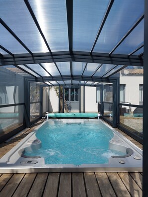 A covered outdoor swimming pool with a wooden deck, glass walls, and a metal roof structure.