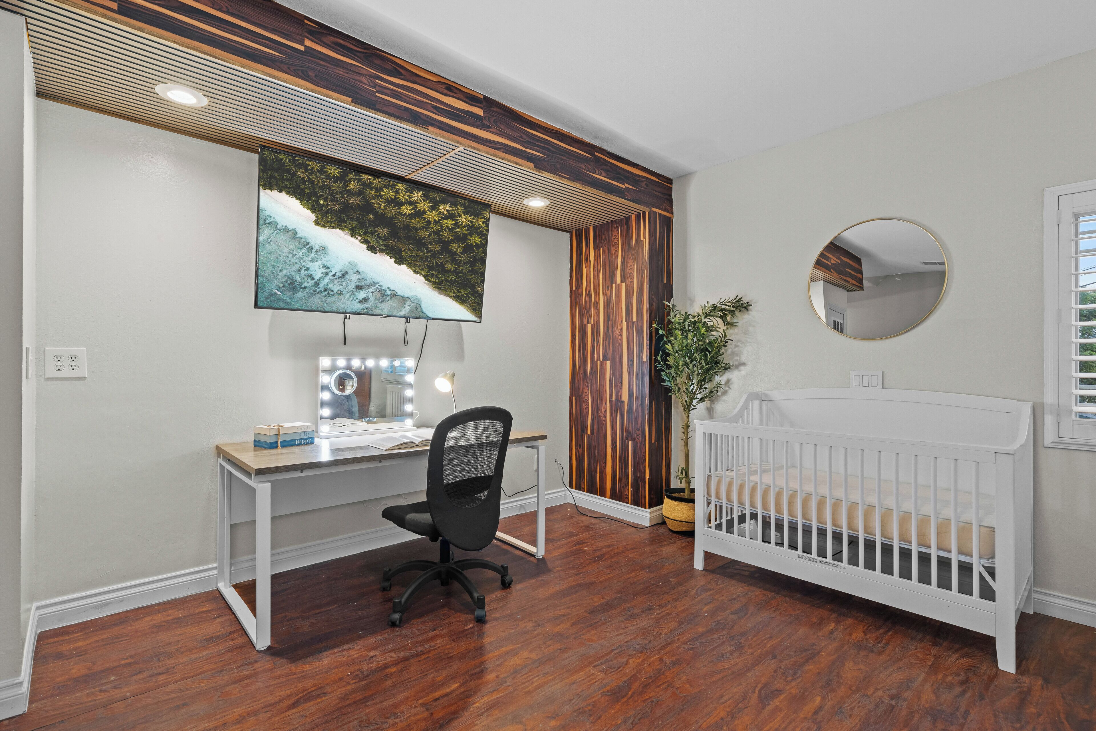 A modern nursery with a wooden floor, a white crib, a desk with a computer, a large TV, and a mirror.