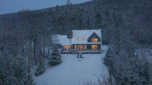 A two-story house with a snow-covered landscape, surrounded by trees.