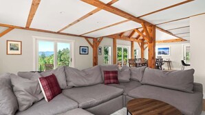 A modern living room with a sectional sofa, wooden beams, and large windows offering a view of greenery.
