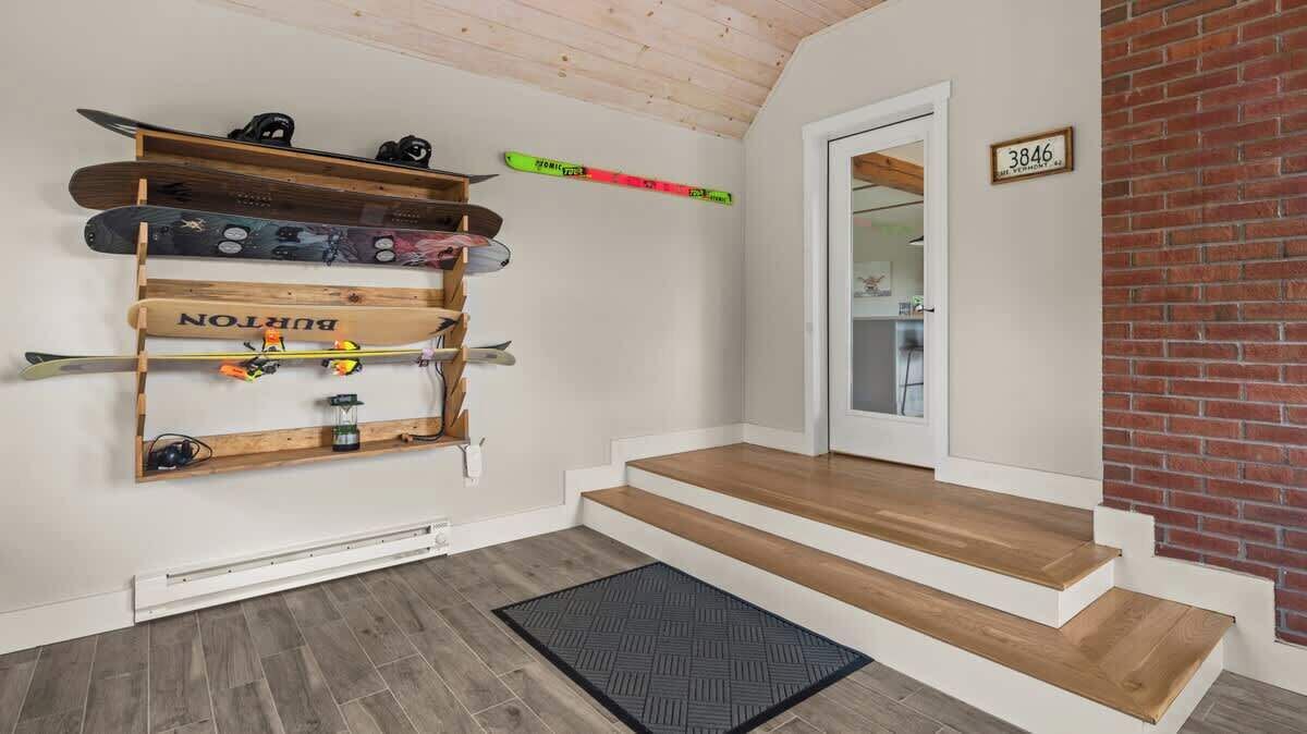 Ein Zimmer mit Holzboden, einer schwarzen Matte am Eingang, einem Skiständer mit Skiern und Snowboards sowie einer Tür mit einer Nummer.'