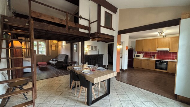 A spacious living area with a high ceiling, exposed wooden beams, a dining table set for four, and a kitchen with wooden cabinets and red tiles. - T3 duplex house, atypical, with wooded garden, very large terrace and parking space (Toulouse)