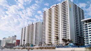 Een hoog hotel met meerdere balkons, een strand op de voorgrond en palmbomen.