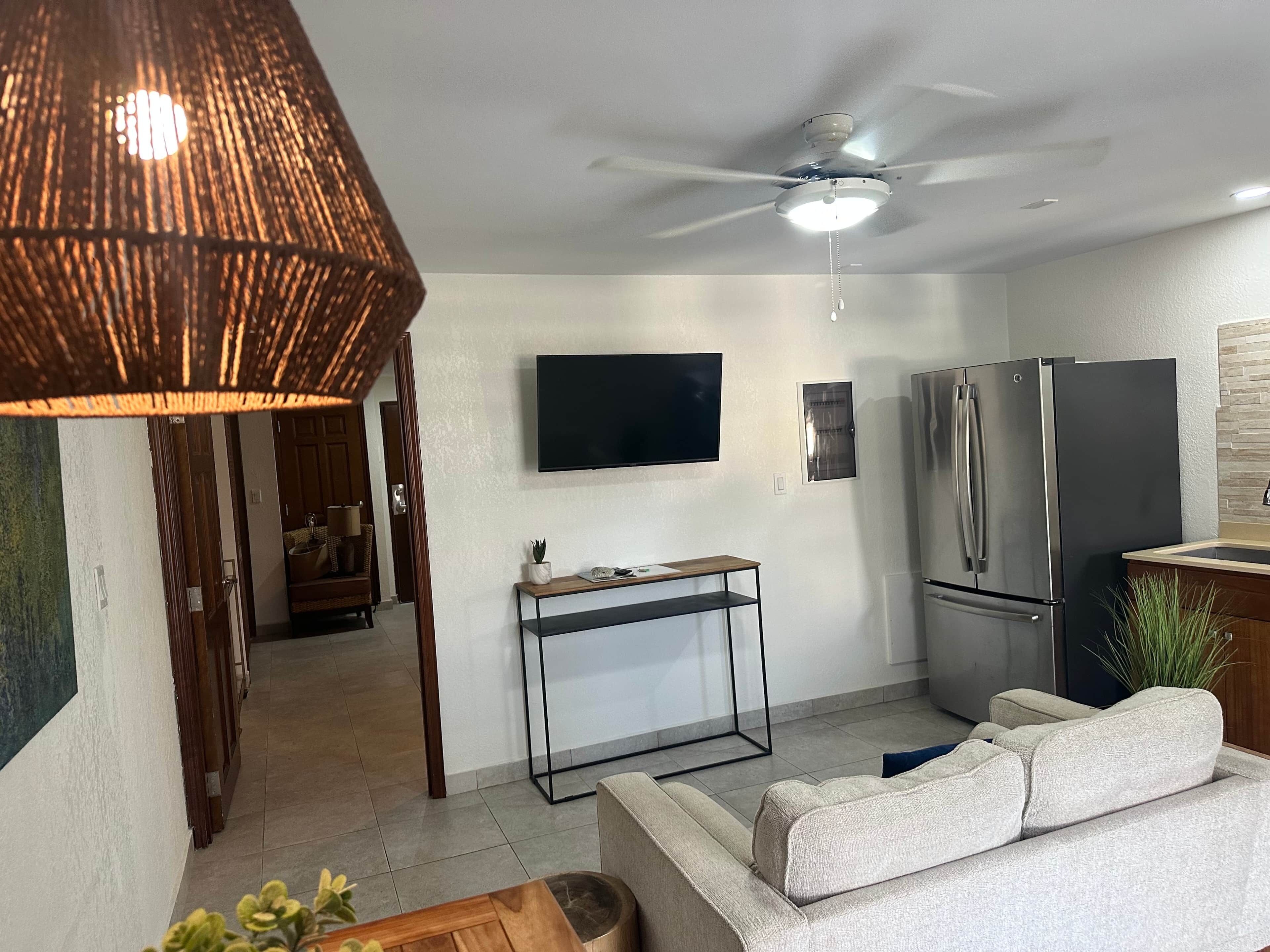 Un salon moderne comprenant un canapé, un téléviseur, un ventilateur de plafond et un coin cuisine avec un réfrigérateur.