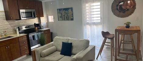 A modern kitchen with wooden cabinets, a stainless steel oven, and a microwave. There is a beige sofa with a blue pillow, a wooden stool, and a round wooden table with a potted plant.