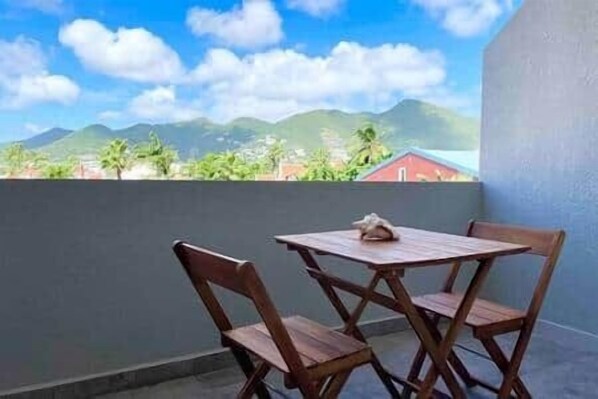 A balcony with a table and chairs, overlooking a mountainous landscape with greenery and buildings.