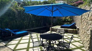 A patio with a table and chairs, a blue umbrella, and lounge chairs by a pool. - Modern apartment with shared pool and generator (2-2B) (Simpson Bay)