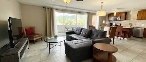Una sala moderna con sofá, mesa de centro y televisor, un balcón con vistas y una zona de cocina con microondas.