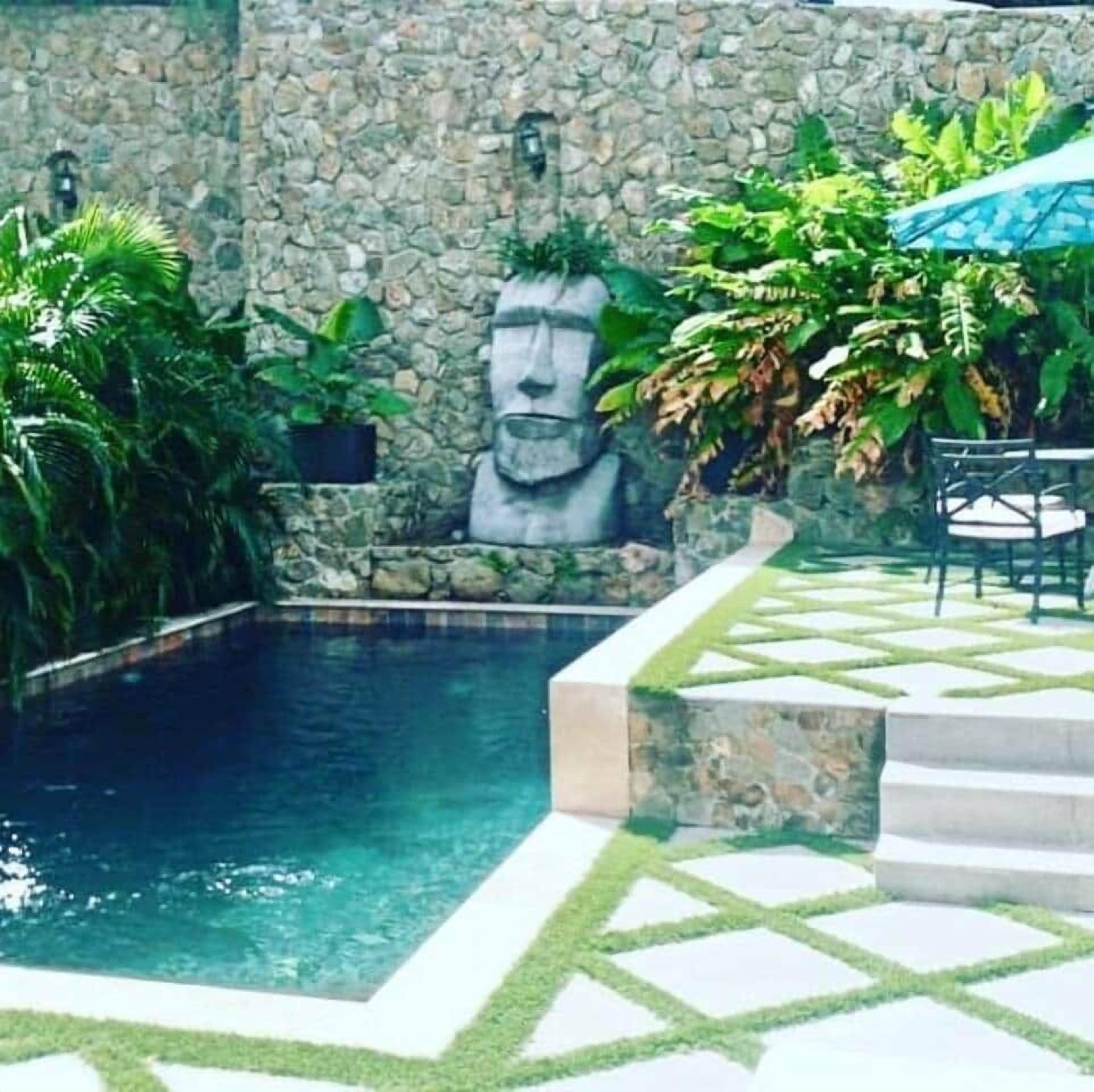 A swimming pool with a stone wall and a statue, surrounded by greenery and a patterned patio area.