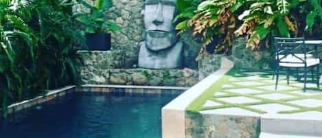 A swimming pool with a stone wall and a statue, surrounded by greenery and a patterned patio area.