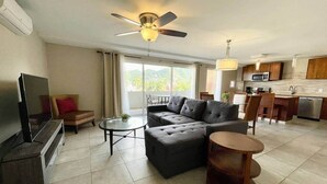 A modern living room with a sofa, coffee table, and TV, a balcony with a view, and a kitchen area with a microwave. - Oasis Apartment in Simpson bay (2-3) (Simpson Bay)