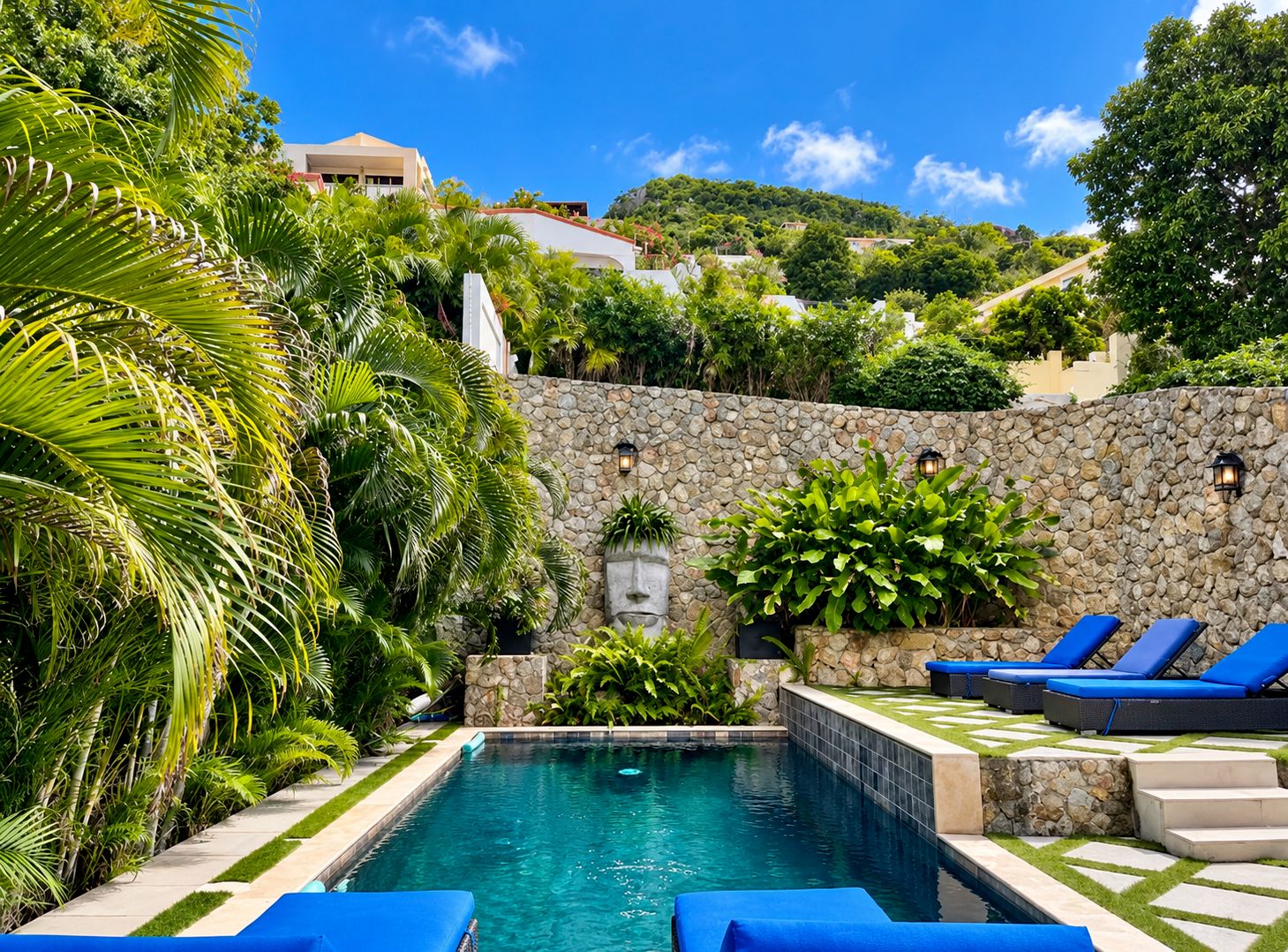 A pool area with stone walls, palm trees, and blue lounge chairs.