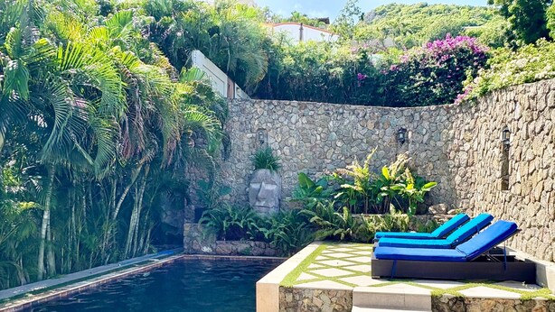 A pool area with stone walls, lush greenery, and blue lounge chairs. - Oasis Apartment in Simpson bay (2-3) (Simpson Bay)