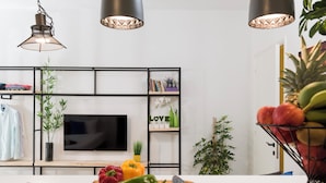 A modern kitchen with a wooden countertop, a cutting board with vegetables, a knife, a TV mounted on the wall, and a bookshelf with decorative items. - Garage Studio in Mar Mikhael (Beirut)