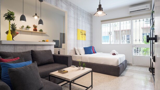A modern living room with a sofa, a coffee table, and a bed. There are pendant lights, a shelf with plants, and a window with a view. - Garage Studio in Mar Mikhael (Beirut)