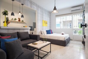 A modern living room with a sofa, a coffee table, and a bed. There are pendant lights, a shelf with plants, and a window with a view. - Garage Studio in Mar Mikhael (Beirut)