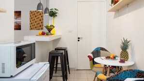 A compact kitchen with a microwave, stools, a small table with fruits, and a door. - Garage Studio in Mar Mikhael (Beirut)
