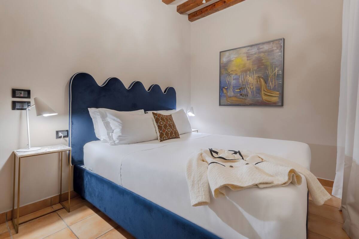 A bedroom with a blue upholstered headboard, white bedding, a bedside lamp, a painting on the wall, and a towel on the bed.