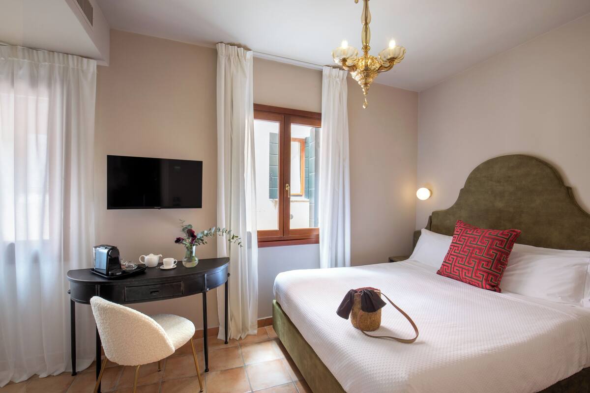 A hotel room with a bed, a desk with a TV, a chair, and a window with curtains.