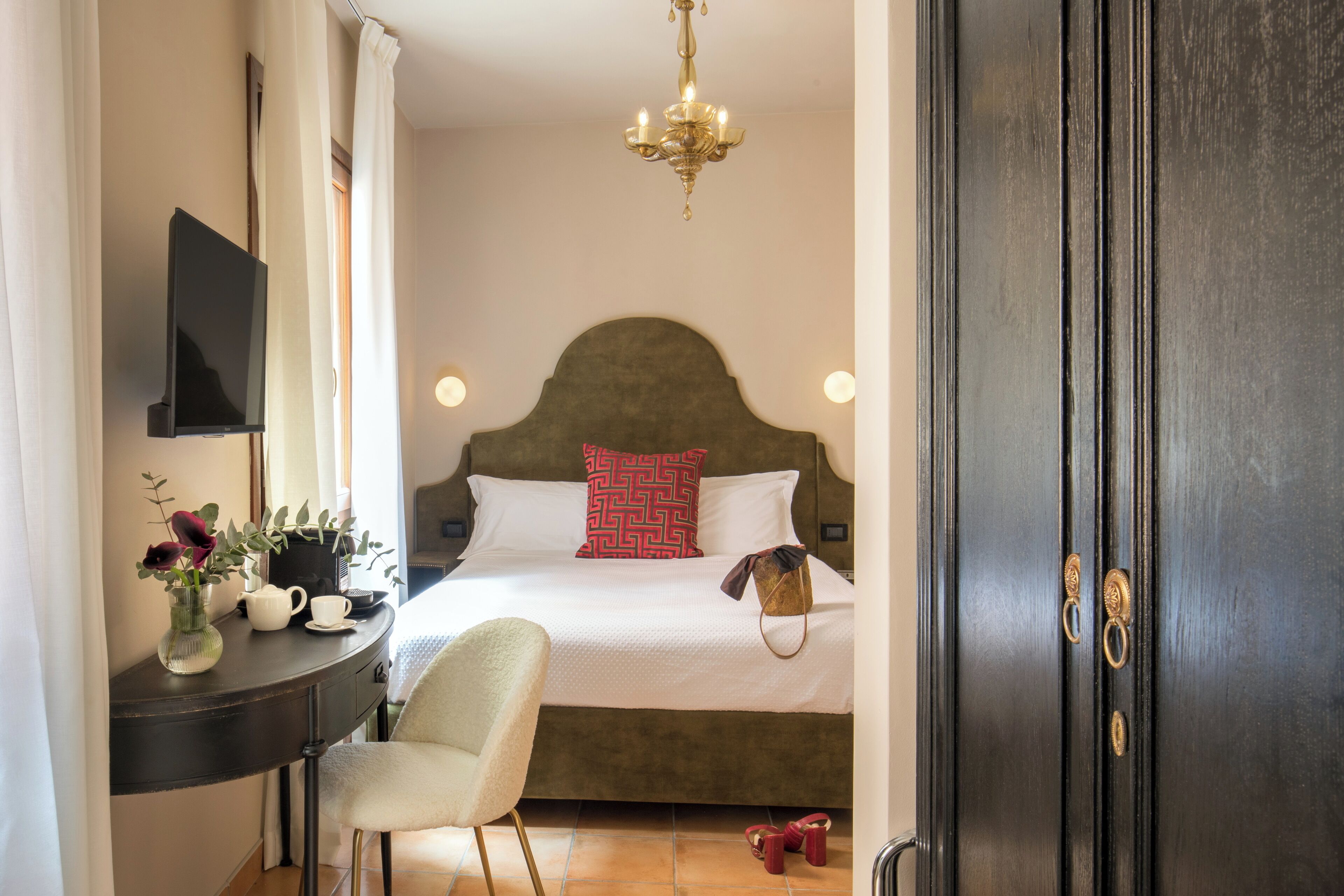 A hotel room with a bed, a desk with a vase of flowers, a chair, and a dark wooden door.
