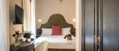 A hotel room with a bed, a desk with a vase of flowers, a chair, and a dark wooden door.