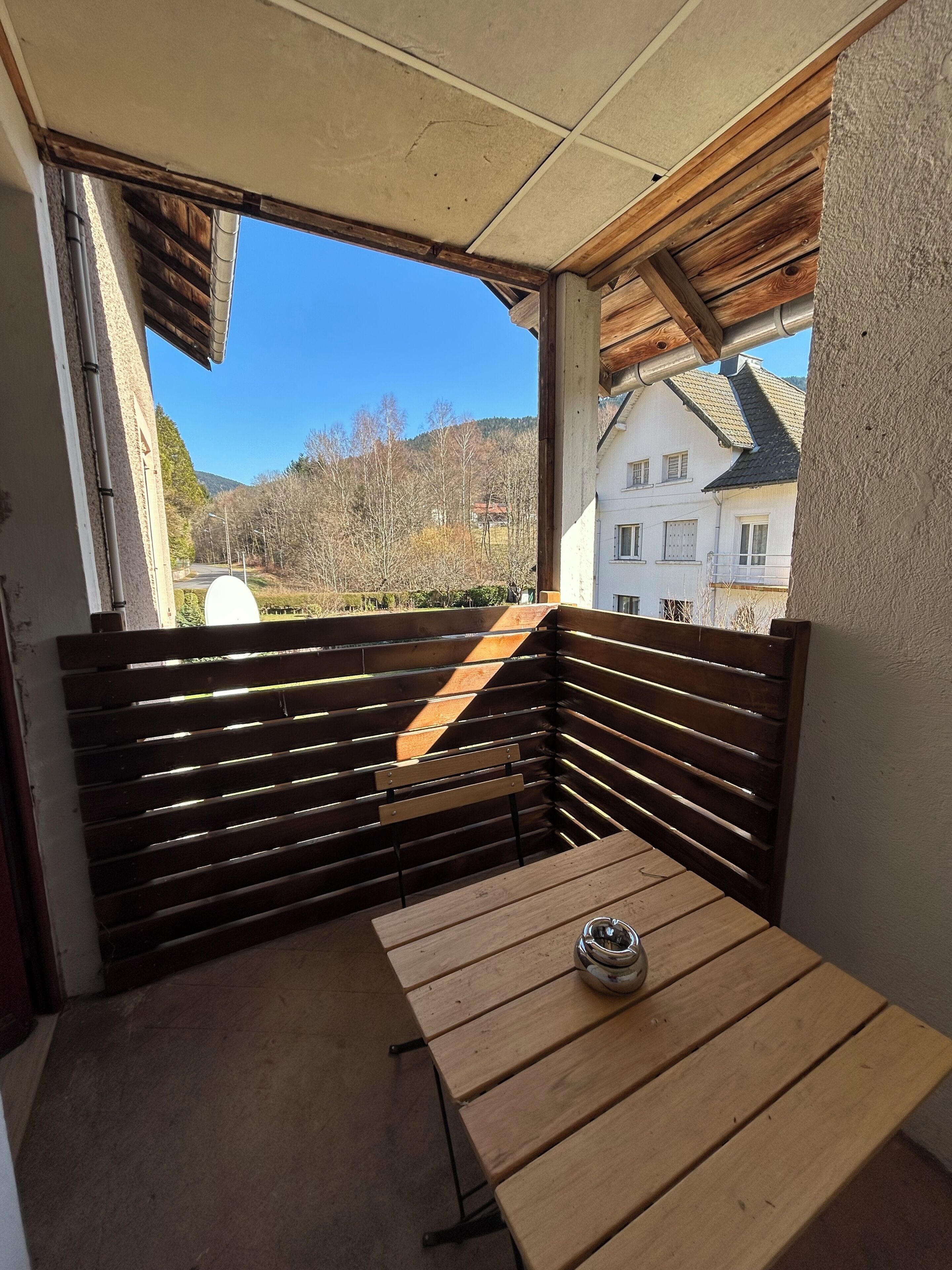 A balcony with a wooden bench and a table, overlooking a residential area and hills.