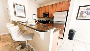 A kitchen with a central island, bar stools, wooden cabinets, stainless steel appliances, and a trash bin. - 5BR Pool+Hot Tub Home Kissimmee Near Disney WDW (Kissimmee)